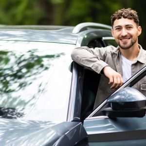 A smiling man standing next to his car. The driver's side door is open, and he is resting his arms on the door frame.