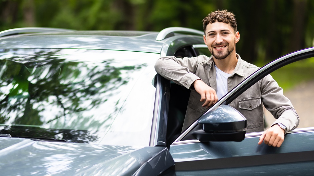 A smiling man standing next to his car. The driver's side door is open, and he is resting his arms on the door frame.