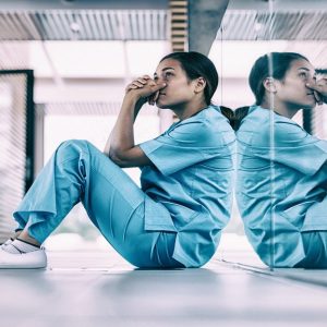 A healthcare working wearing scrubs sits with their back to a mirror with their hands clasped near their face.
