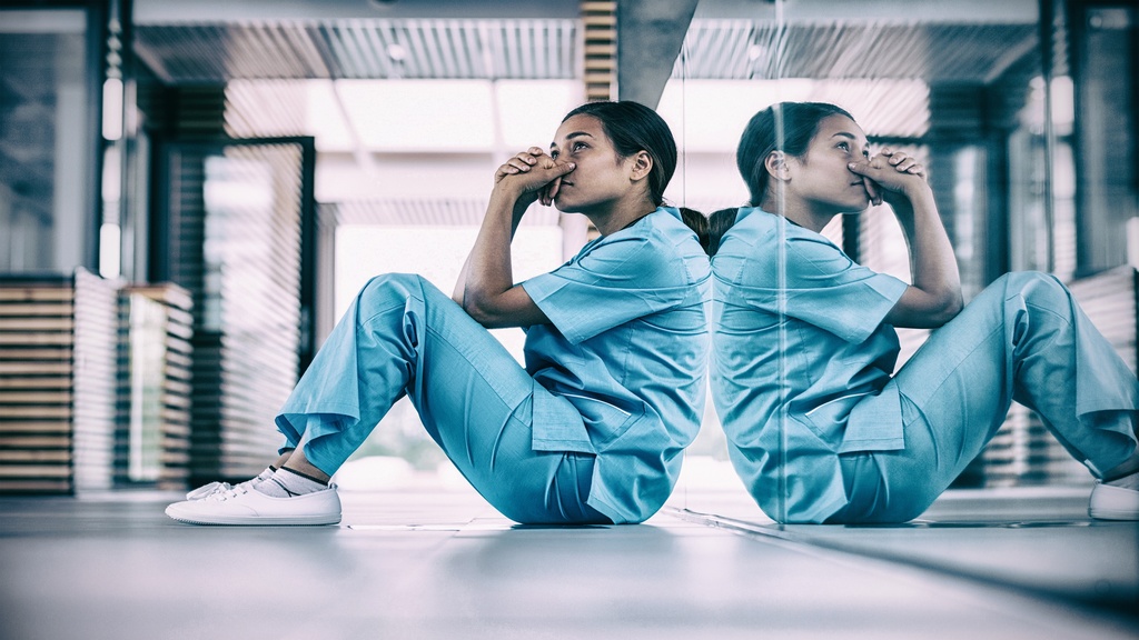 A healthcare working wearing scrubs sits with their back to a mirror with their hands clasped near their face.
