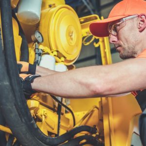 A man crouched next to an excavator engine he's working on. He's wearing orange reflective clothing and protective glasses.