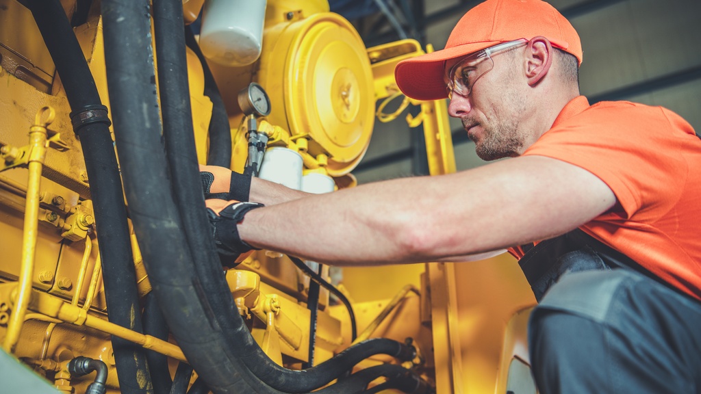 A man crouched next to an excavator engine he's working on. He's wearing orange reflective clothing and protective glasses.