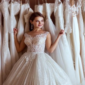 A young woman wearing a wedding dress smiles while leaning towards a clothing rack with multiple wedding gowns.