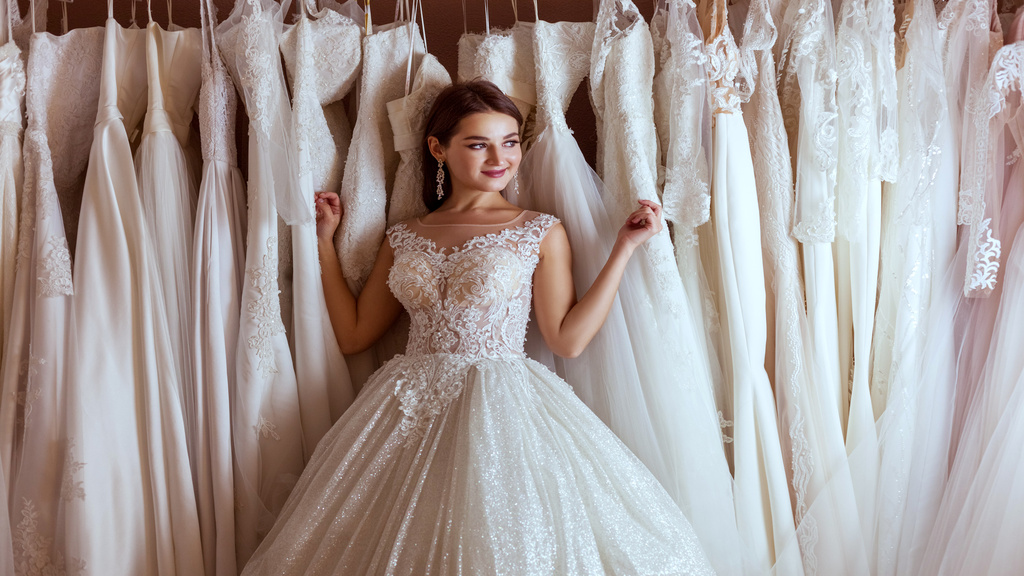 A young woman wearing a wedding dress smiles while leaning towards a clothing rack with multiple wedding gowns.