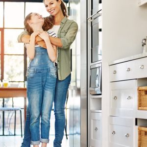 A mother hugging her tween daughter from behind in the kitchen. The daughter smiles and cranes her head back to look at her mom.