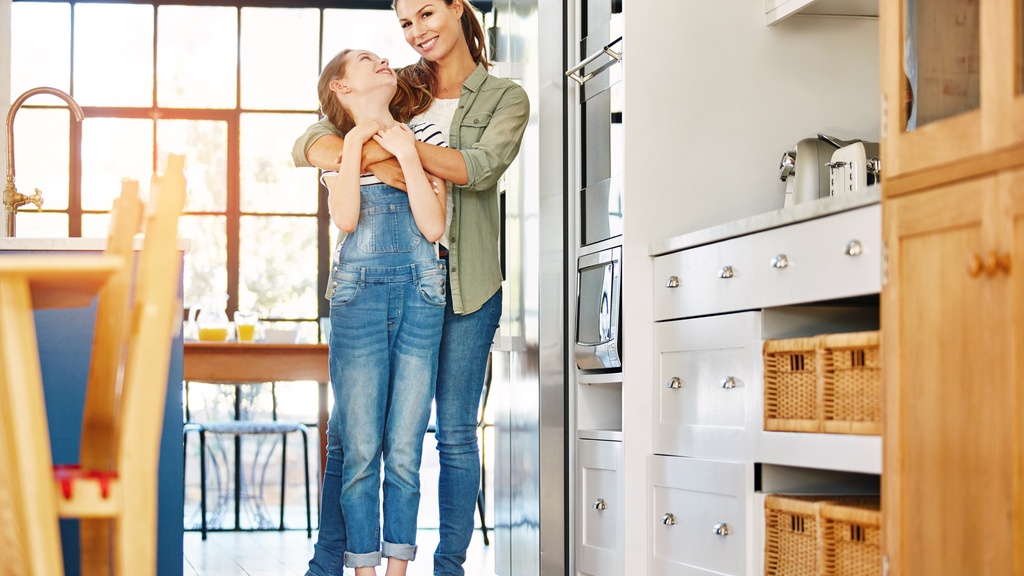 A mother hugging her tween daughter from behind in the kitchen. The daughter smiles and cranes her head back to look at her mom.