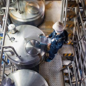 An industrial plant worker wearing a hard hat holds a clipboard while inspecting steel tanks at a processing plant.