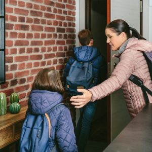 A woman wearing a heavy jacket, a backpack, and a walking stick, leading two kids out the door from inside a room.