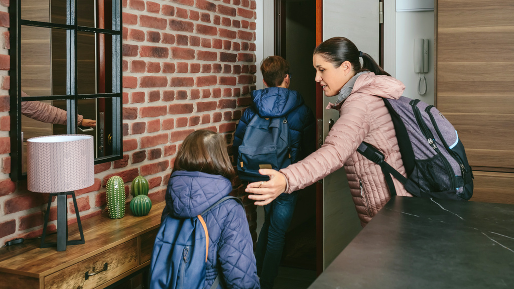 A woman wearing a heavy jacket, a backpack, and a walking stick, leading two kids out the door from inside a room.