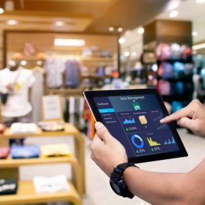A manager holding a tablet in front of a store display. The tablet shows graphs and statistics for the business.