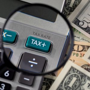 A calculator with a button that says "TAX+" on it, sitting on some money. There is a magnifying glass over the button.