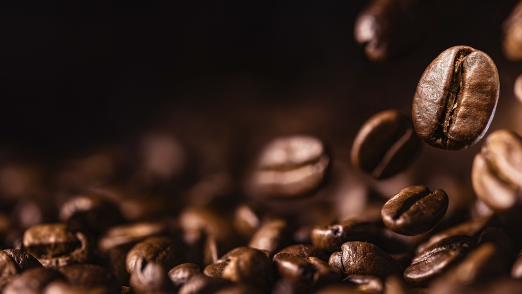 A close-up of coffee beans falling onto a table. The background is brown, and the beans are whole and intact.