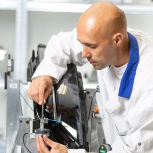 Technician in white lab coat calibrates a precision gauge on a metal fixture inside a bright industrial inspection lab.