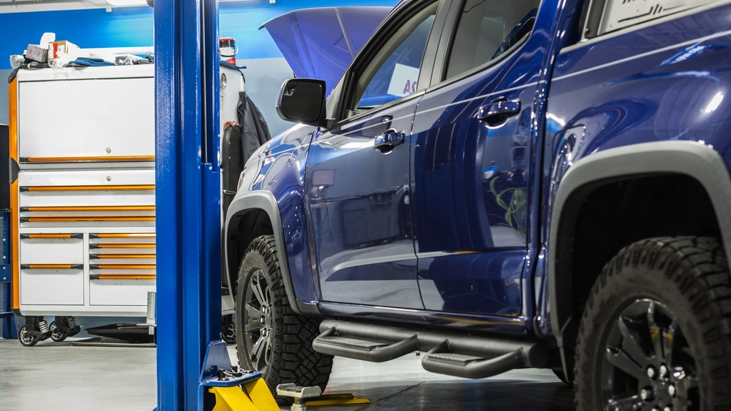 A rear and side view of a blue pickup truck with black rims in a mechanic shop with the engine hood open.