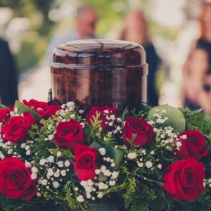 A polished wooden cremation urn surrounded by red roses on a table, with blurred mourners in the background.