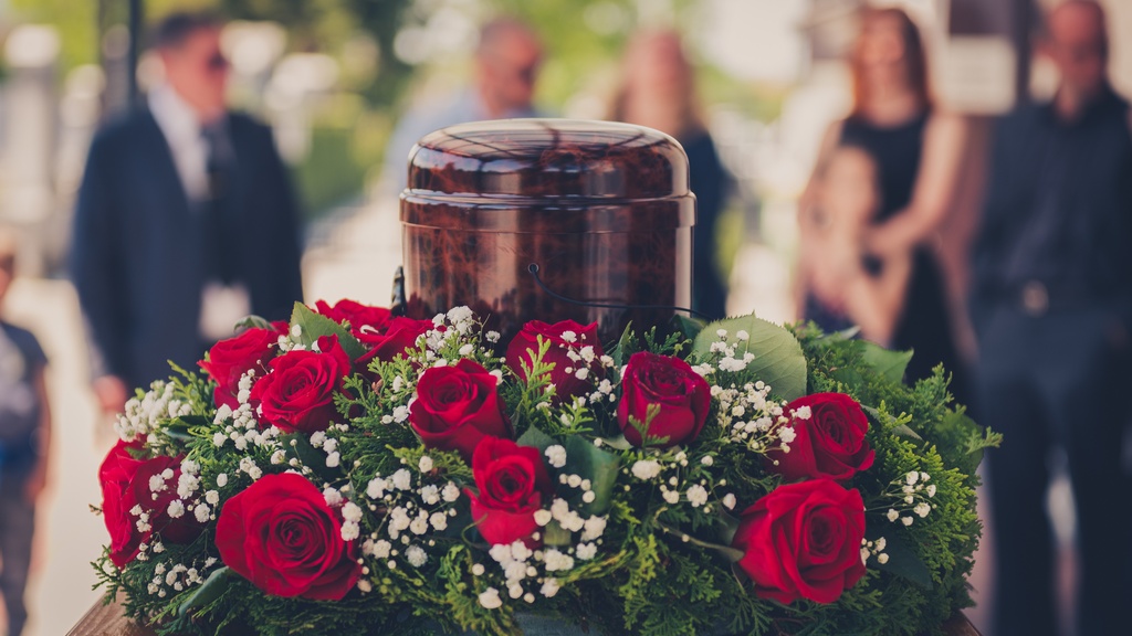 A polished wooden cremation urn surrounded by red roses on a table, with blurred mourners in the background.
