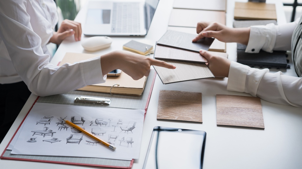 An interior designer sits across a homeowner as they collaborate over samples, swatches, and floor plans on the desk.