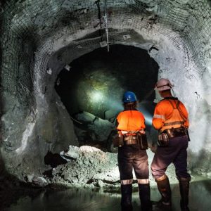Two workers wearing bright orange vests and helmets, using a flashlight to illuminate a path inside a mine tunnel.