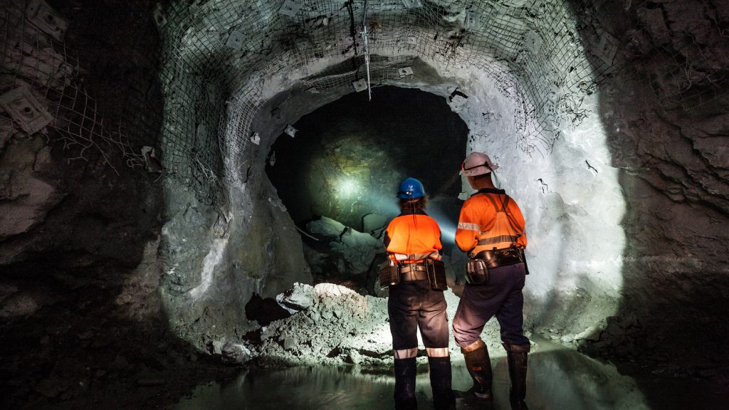 Two workers wearing bright orange vests and helmets, using a flashlight to illuminate a path inside a mine tunnel.