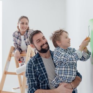 A mother stands on a ladder and watches as a father holds his son, who paints a wall green with a roller.
