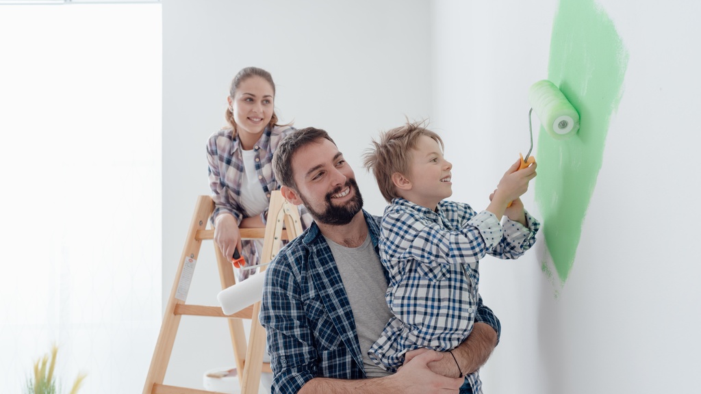 A mother stands on a ladder and watches as a father holds his son, who paints a wall green with a roller.