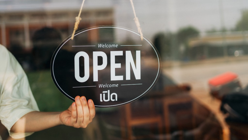 A hand reaches over behind a business's window and flips a hanging sign so it reads the words "Welcome" and "Open."