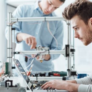 A man is sitting at a desk and is typing on a computer. There is a man behind him fiddling with a 3D printer.