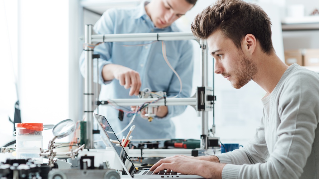 A man is sitting at a desk and is typing on a computer. There is a man behind him fiddling with a 3D printer.