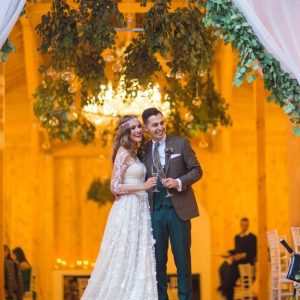 A man in a suit and a woman in a white dress are standing next to each other. Soft white curtains are draped above them.