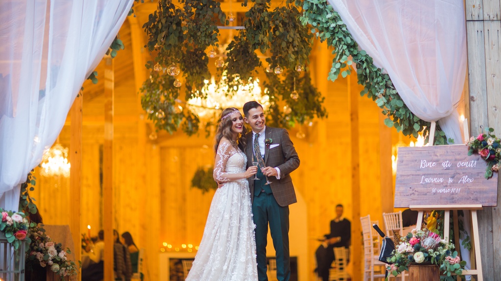 A man in a suit and a woman in a white dress are standing next to each other. Soft white curtains are draped above them.