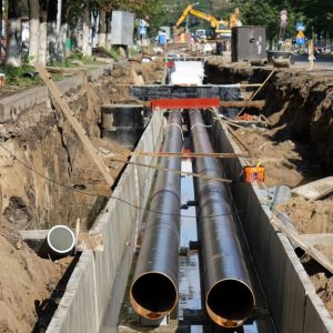 A ditch is dug out along a city street. There are two long pipes running parallel with each other that are uncovered.