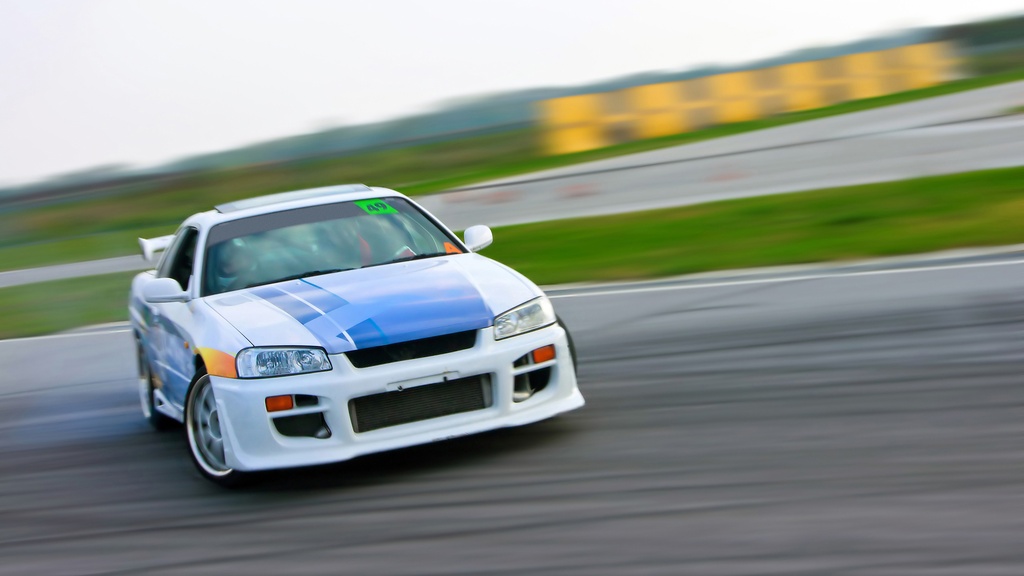 A blue and white race car drifts on a race track. The green grass lies behind it, along with another roadway.