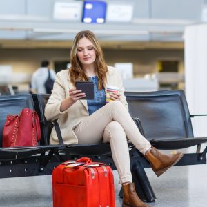 A woman sits in an airport with a red suitcase and a red handbag. She holds a to-go cup and reads on a digital device.