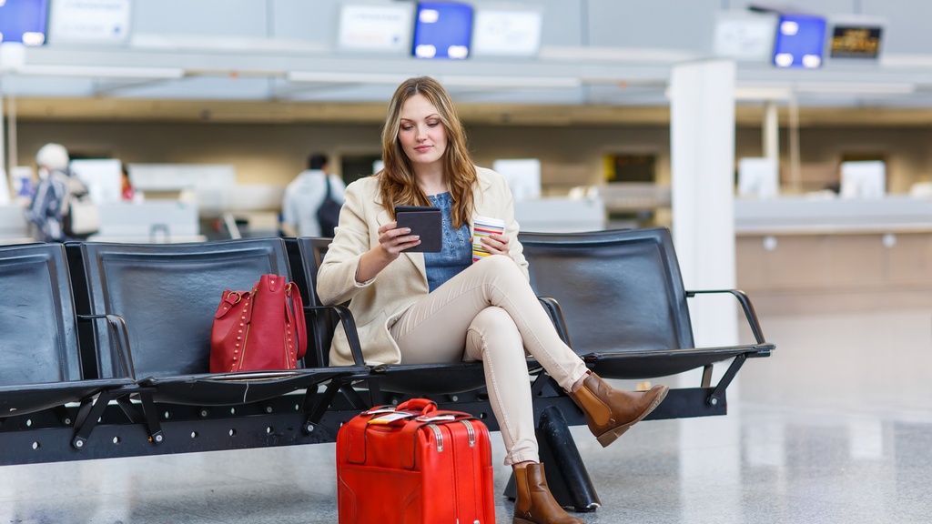 A woman sits in an airport with a red suitcase and a red handbag. She holds a to-go cup and reads on a digital device.