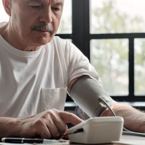An older man in a white t-shirt checks his blood pressure with an automatic monitor while seated at a table near a window.