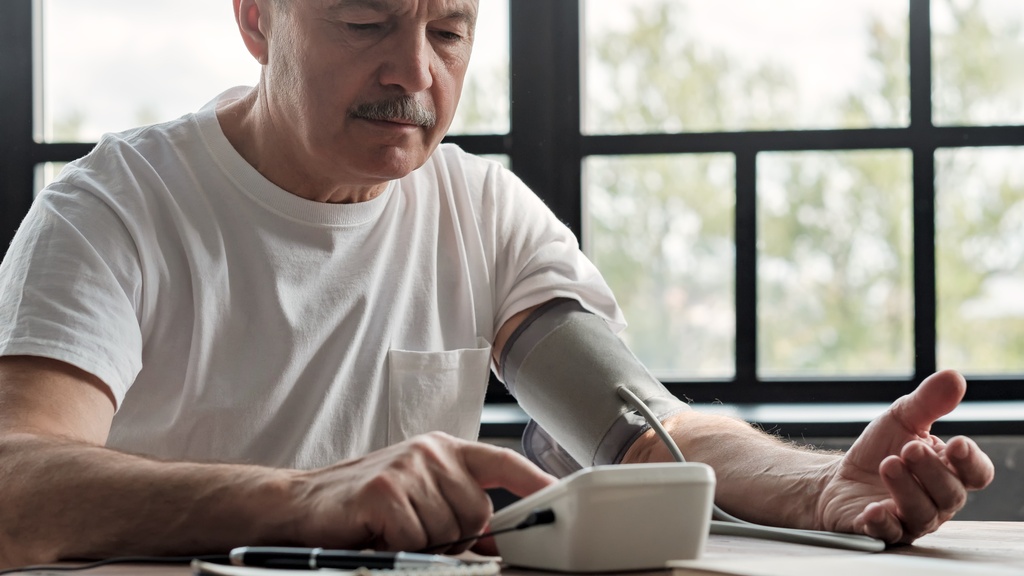 An older man in a white t-shirt checks his blood pressure with an automatic monitor while seated at a table near a window.