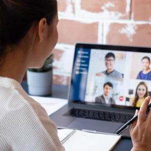 A young woman sits at her remote desk as she waves to the screen. The screen features a team meeting with four people.