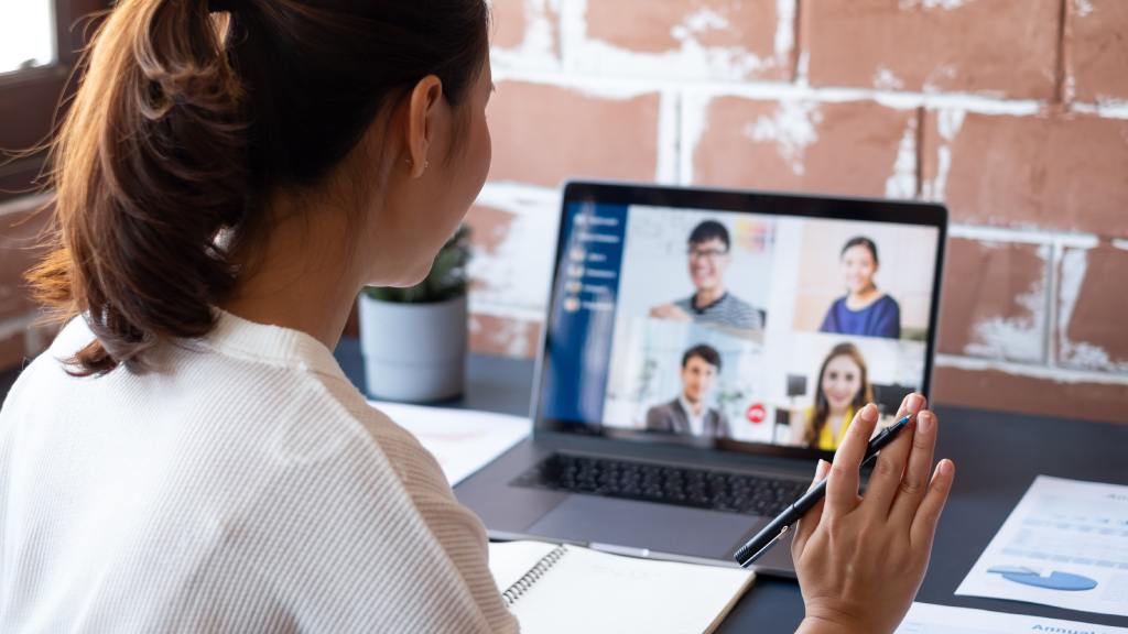 A young woman sits at her remote desk as she waves to the screen. The screen features a team meeting with four people.