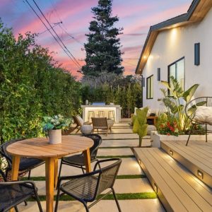 The backyard patio of a home that is well lit and has plenty of seating options. There is a large hedge enclosing the space.