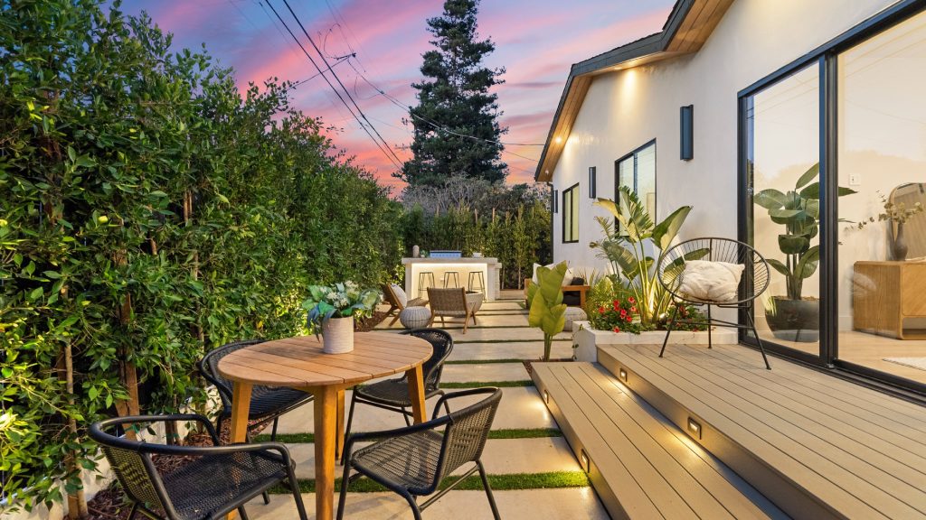 The backyard patio of a home that is well lit and has plenty of seating options. There is a large hedge enclosing the space.
