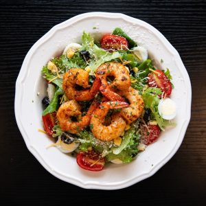A white plate of seasoned grilled shrimp over a fresh salad with cherry tomatoes, olives, and hard-boiled eggs on a dark wood table.