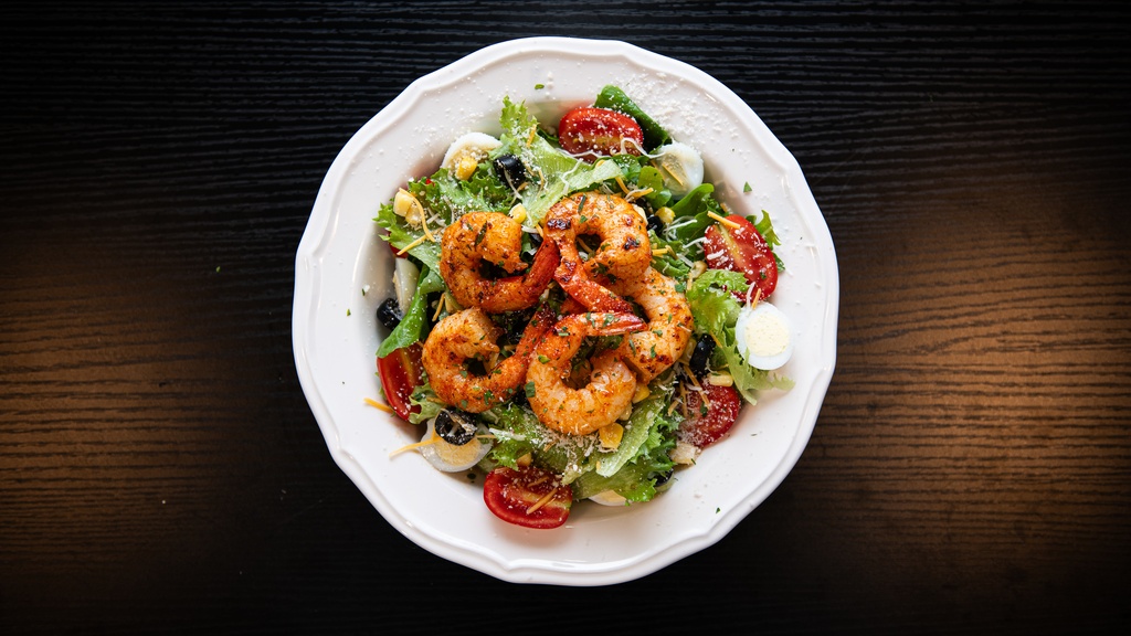 A white plate of seasoned grilled shrimp over a fresh salad with cherry tomatoes, olives, and hard-boiled eggs on a dark wood table.