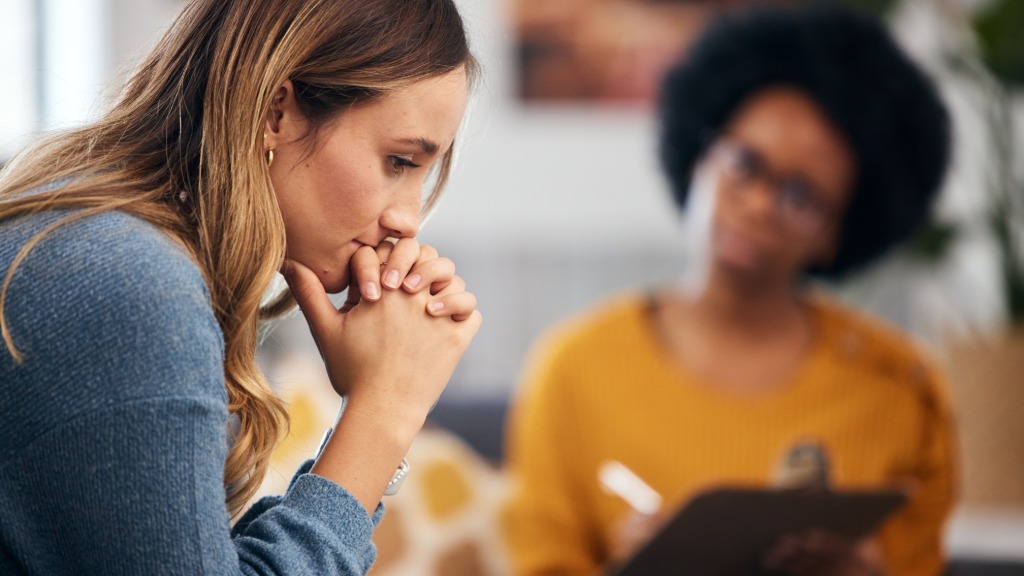 A woman sits with her hands clasped, looking down during a therapy session as a therapist takes notes in the background.