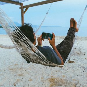 man laying in a hammack on the beach using his phone playing games