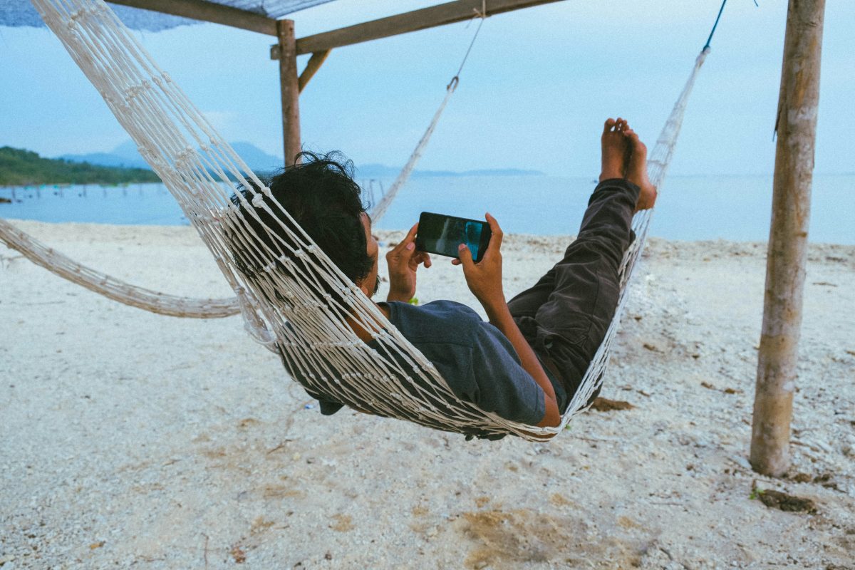 man laying in a hammack on the beach using his phone playing games