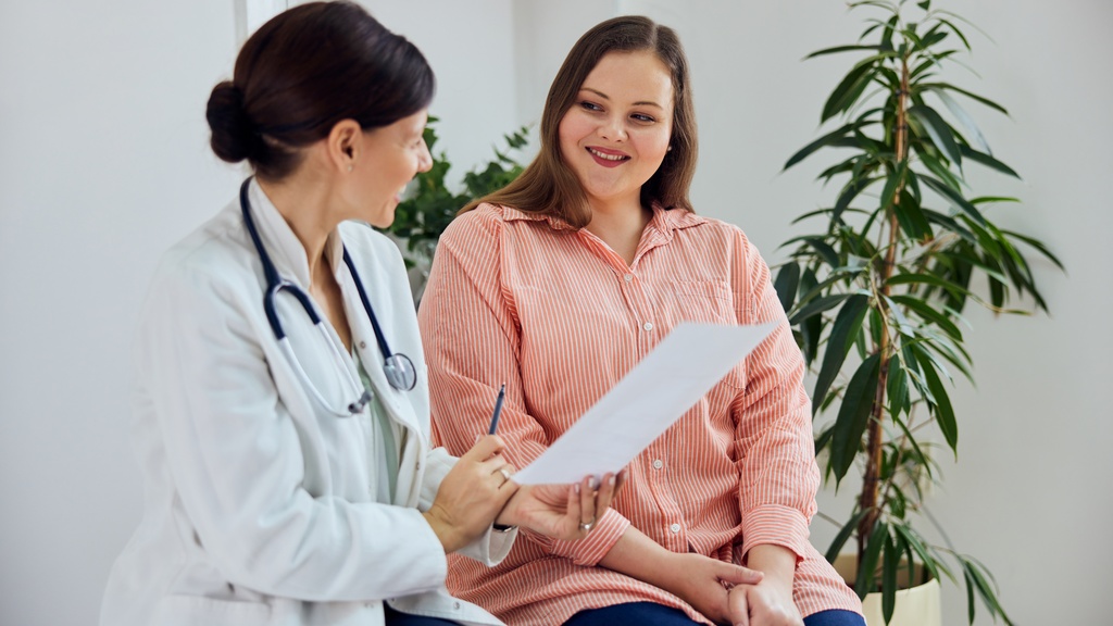 A woman in a doctor's coat with a stethoscope around their neck talking to a woman in casual attire sitting on a chair.