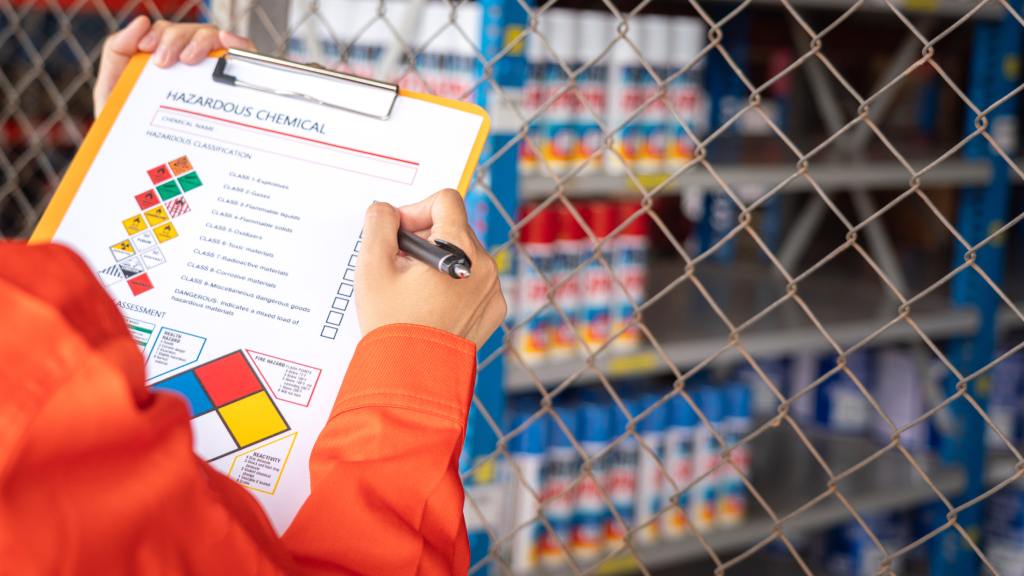 A safety worker in an orange jacket reviews a hazardous chemical classification checklist in a chemical storage facility.