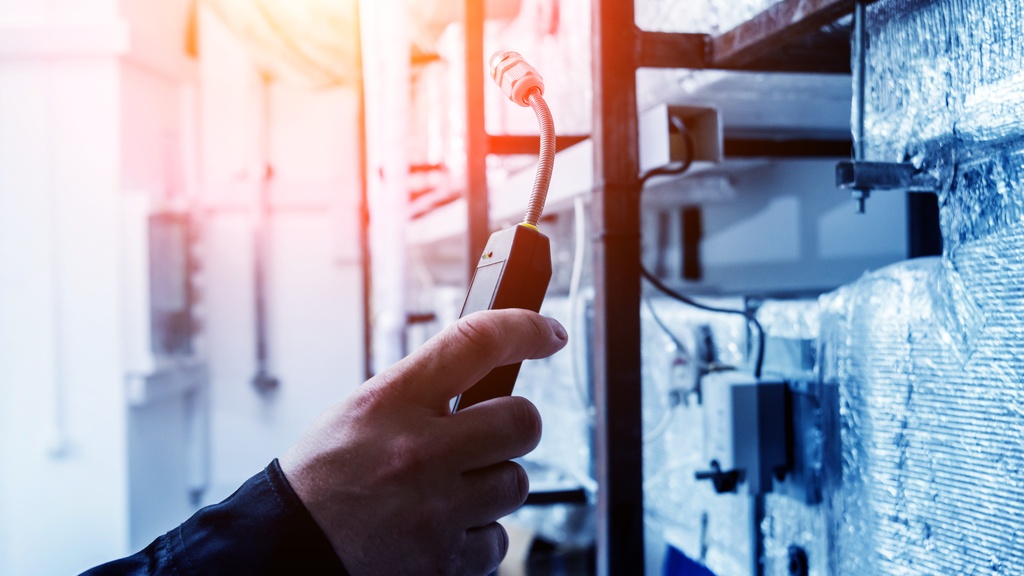 A technician uses a handheld electronic leak detector near insulated industrial HVAC equipment and wiring in a facility.