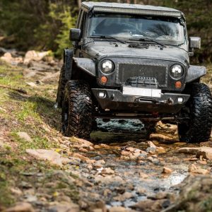 A rugged black Jeep Wrangler equipped with oversized tires and a winch tackles a rocky, wet forest trail.