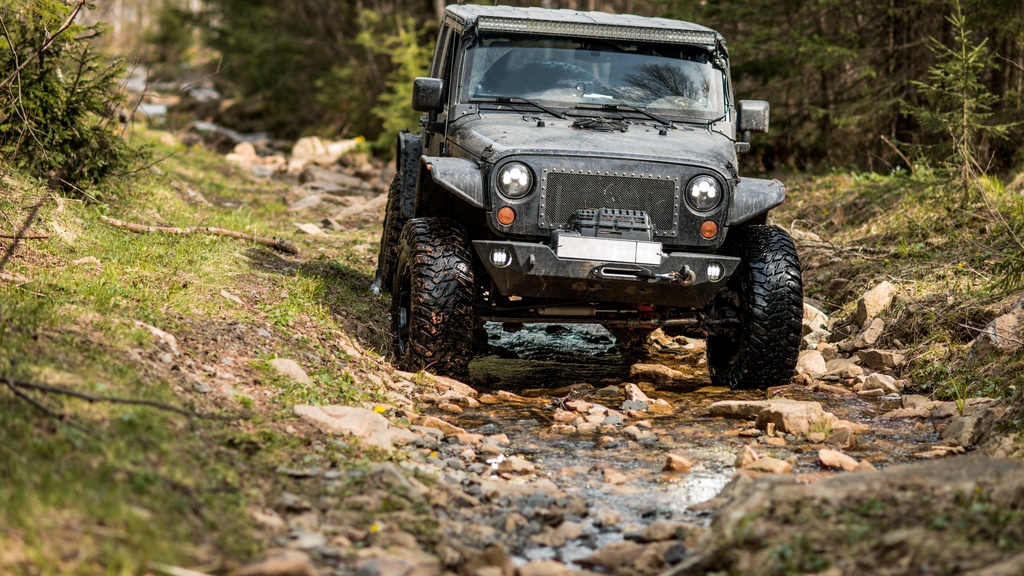 A rugged black Jeep Wrangler equipped with oversized tires and a winch tackles a rocky, wet forest trail.